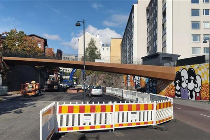 En rostfärgad bro över gatan. På gatan pågår vägarbete, vid kanten finns klippor, höghus och träd.