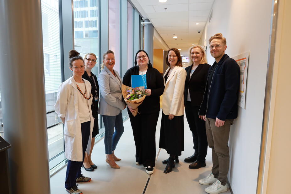 En grupp personer står i en korridor. Personen i mitten håller en blombukett och en blå mapp, medan de andra står bredvid. I bakgrunden syns fönster och en korridor.