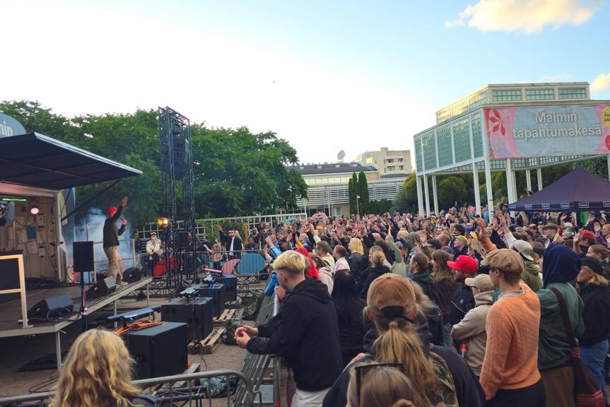 An audience has gathered in front of a stage in Ala-Malmi Park. Malmitalo and a pavilion can be seen in the background. 