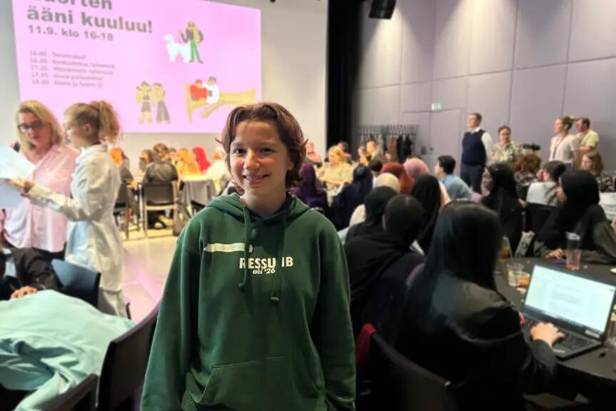 Nuori katsoo kameraan hymyillen isossa salissa, taustalla ihmisiä istuu pöydissä. Takaseinälle on heijastettu esitys, jossa lukee Nuorten ääni kuuluu. 